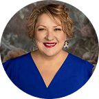 A smiling woman with short curly blonde hair, wearing a royal blue V-neck top and statement earrings, poses in front of a softly blurred floral background.