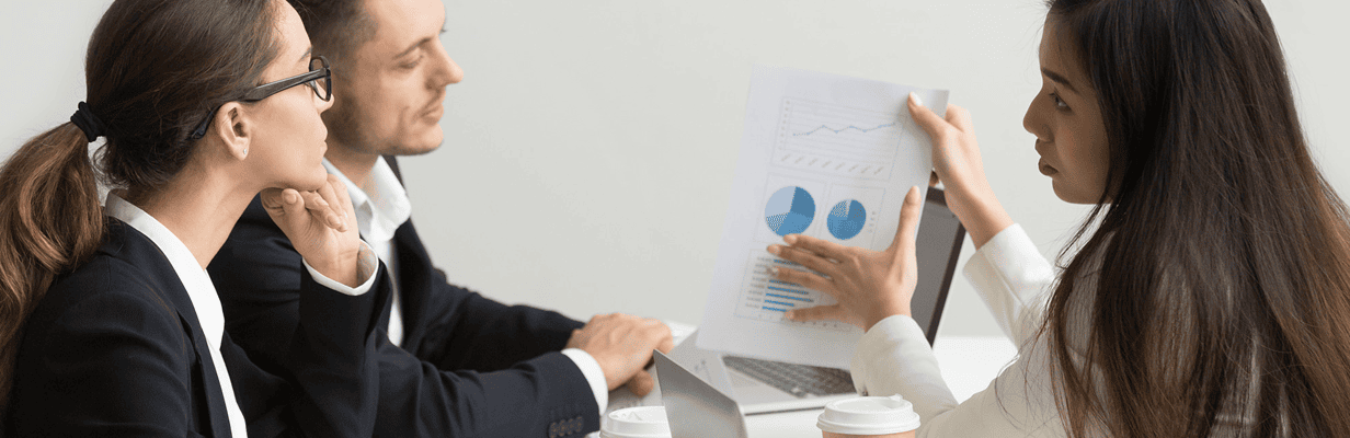 Three business professionals sit at a table. One woman is showing a paper with charts and graphs to two colleagues, who listen attentively. A laptop and coffee cups are on the table.